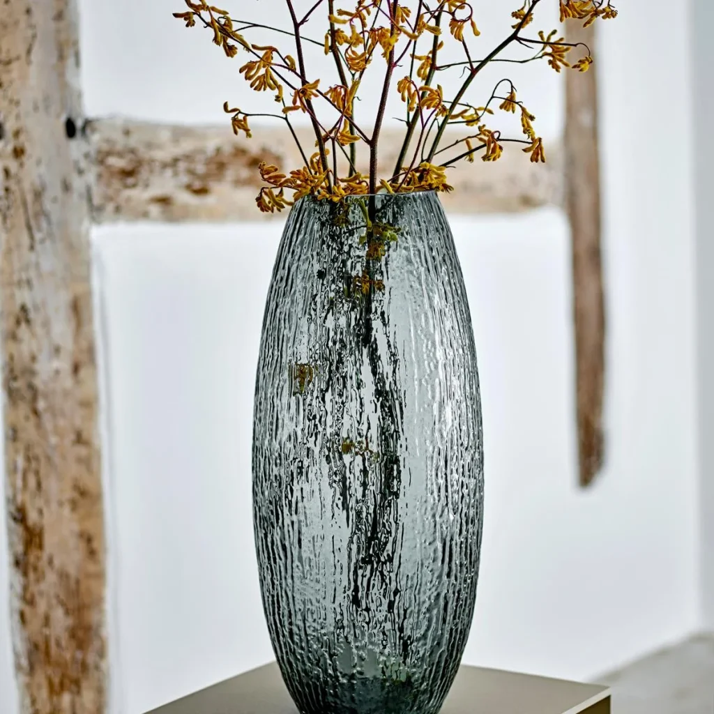 Bodenvase Brock aus grauem Rauchglas mit gelb blühenden Zweigen vor einer hellen Wand mit Holzbalken